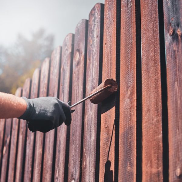 A person is painting a picket fence with a roller brush, applying a rich brown coat.