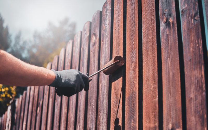 A person is painting a picket fence with a roller brush, applying a rich brown coat.